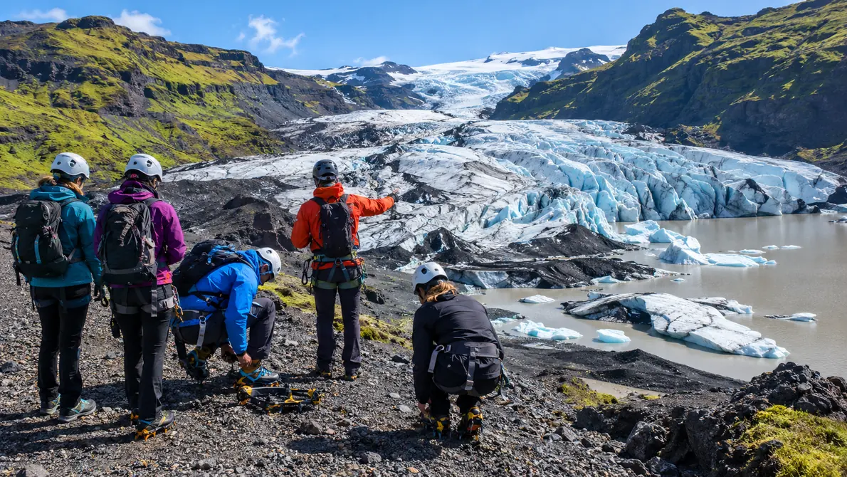 A guide kneeling to fit crampons onto a hiker's boots, a group waiting in helmets, seen from behind