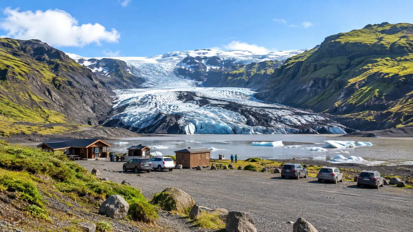 Gravel car park with low base-camp huts, the glacier tongue beyond a ridge of moraine