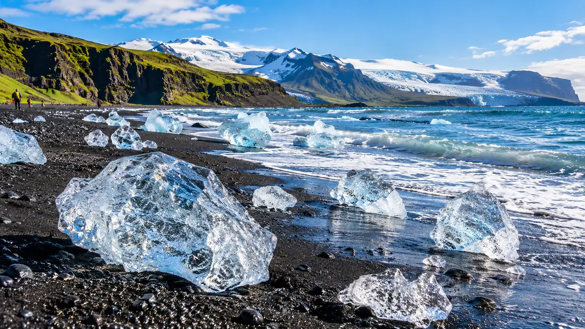 Clear chunks of glacier ice scattered on a black sand beach with waves behind