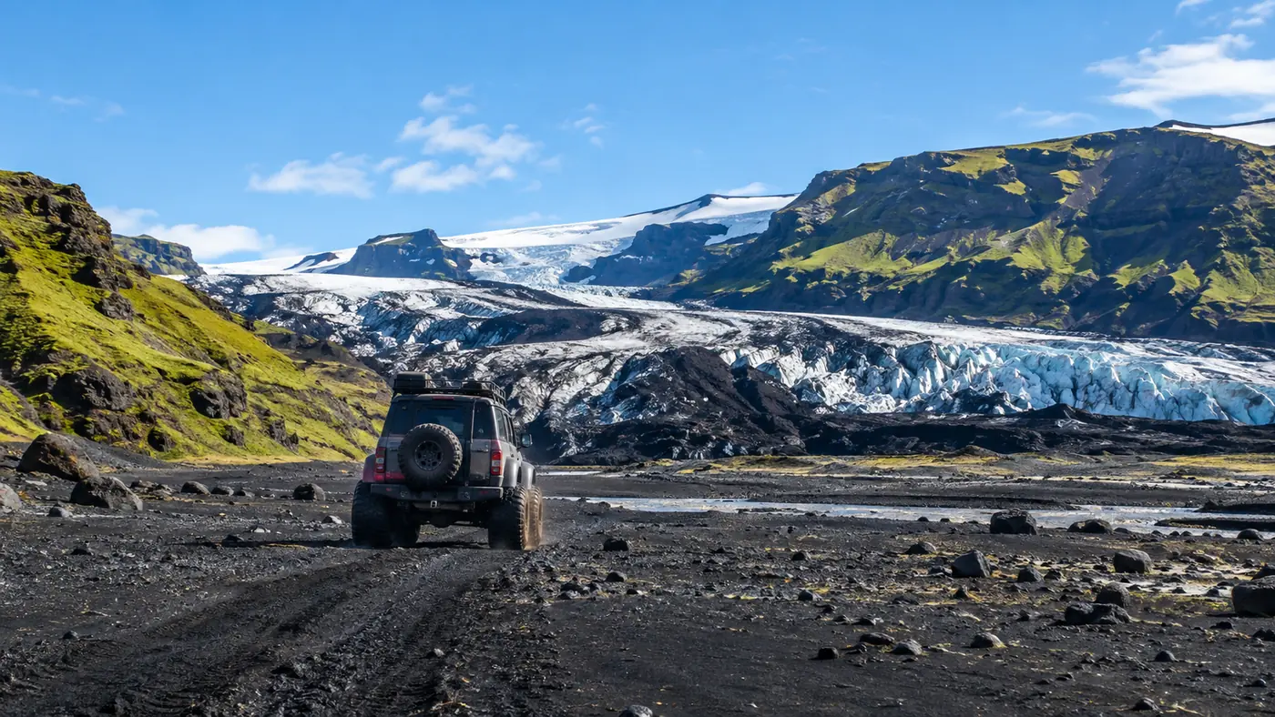 A super jeep with oversized tyres parked on a black sand plain below a glacier